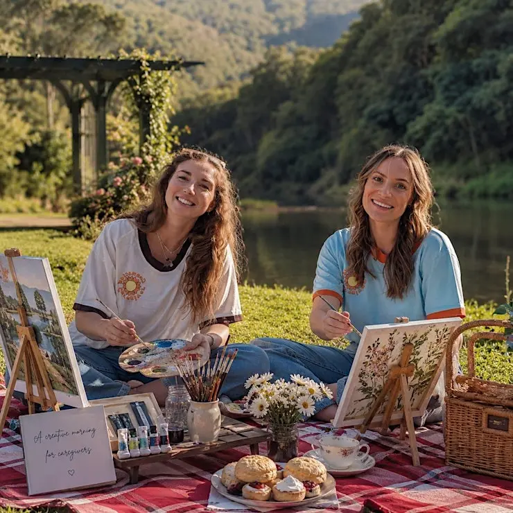 Two friends painting and sharing scones on a riverside picnic rug at Historic Rivermill