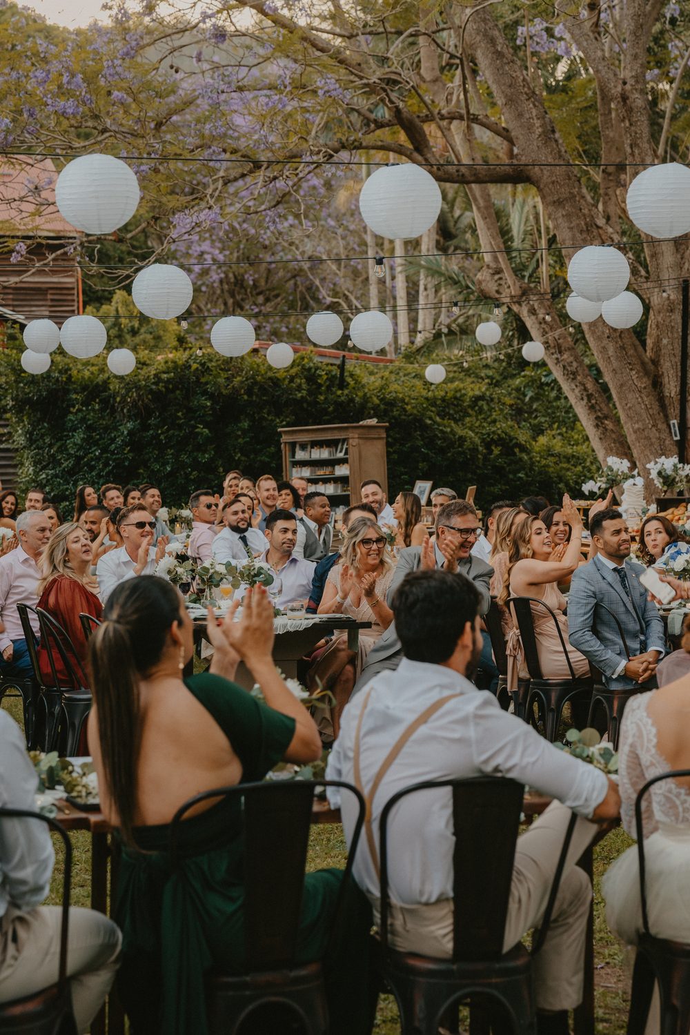 Guests applauding under paper lanterns among the jacarandas