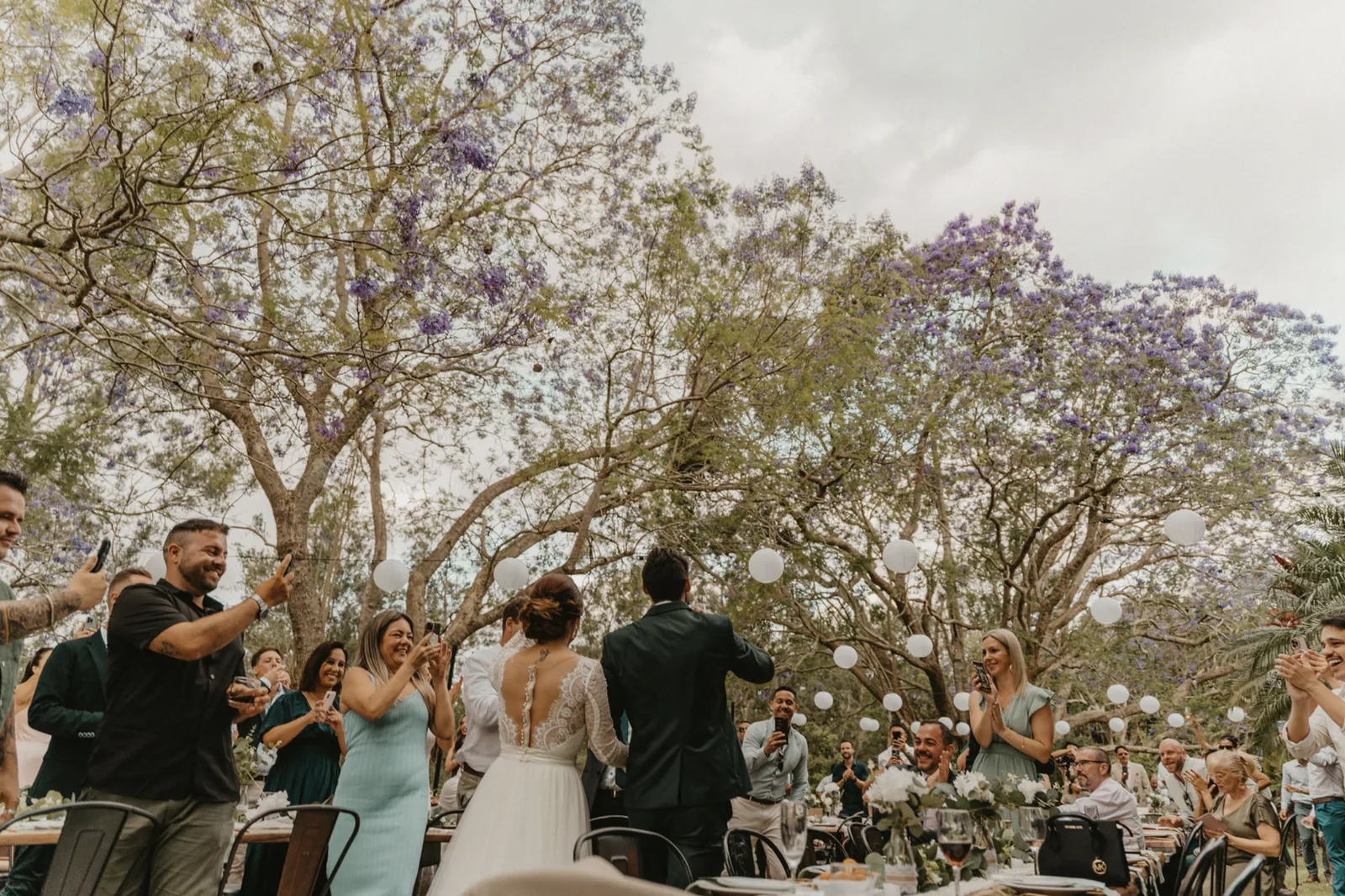 A bride seated at a long white table beneath a jacaranda, petals carpeting the lawn