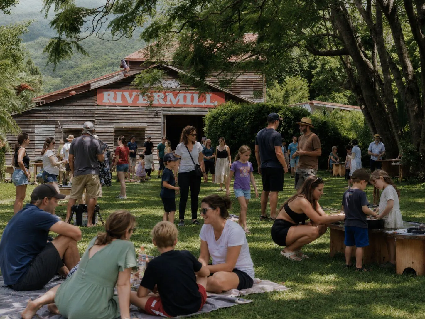 Families gathered on the lawns at Historic Rivermill for an event, with children playing on picnic rugs in front of the red timber barn