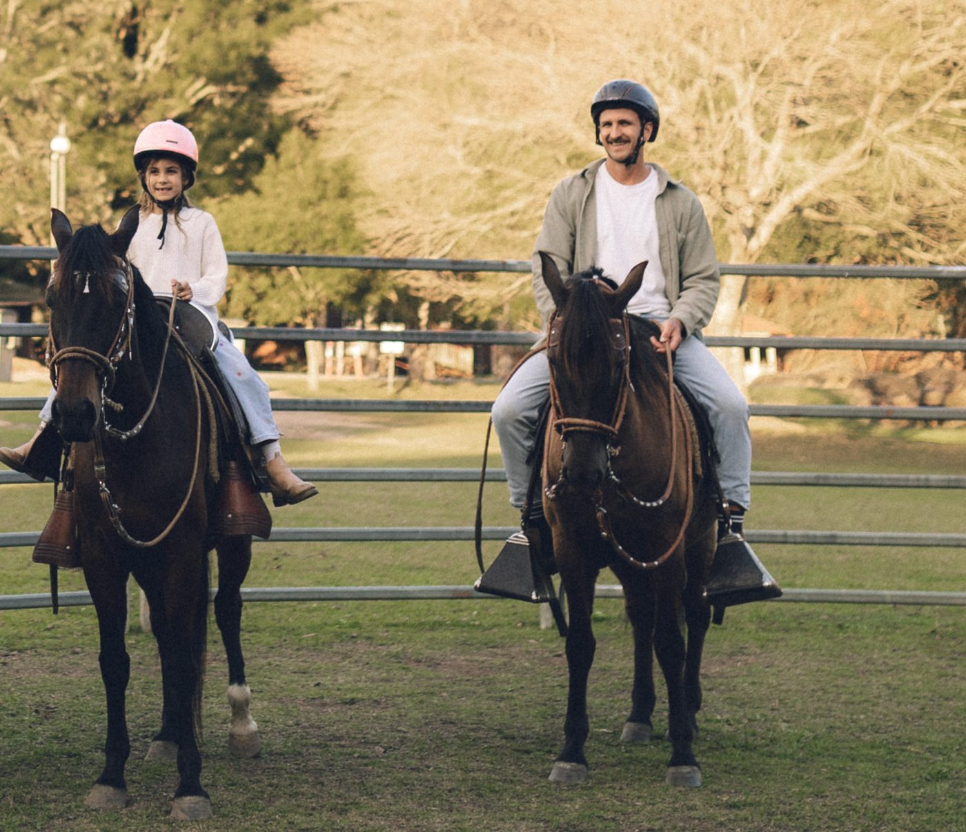 A girl and a man riding side by side on horseback in the paddock at Historic Rivermill, both smiling in the evening light