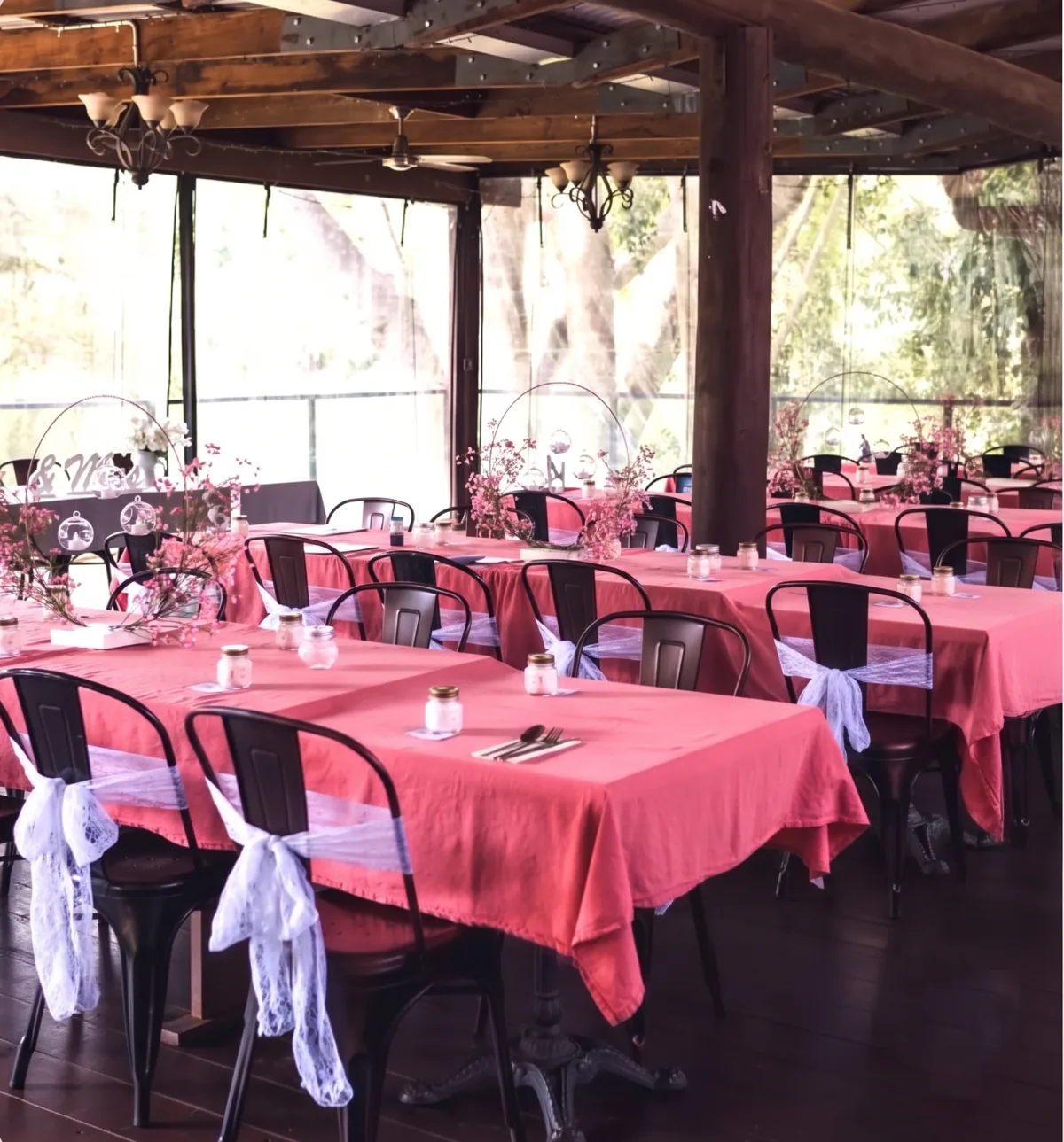 Long tables dressed in coral linen with lace chair sashes and pink blossom on the covered riverside deck, set for a private event