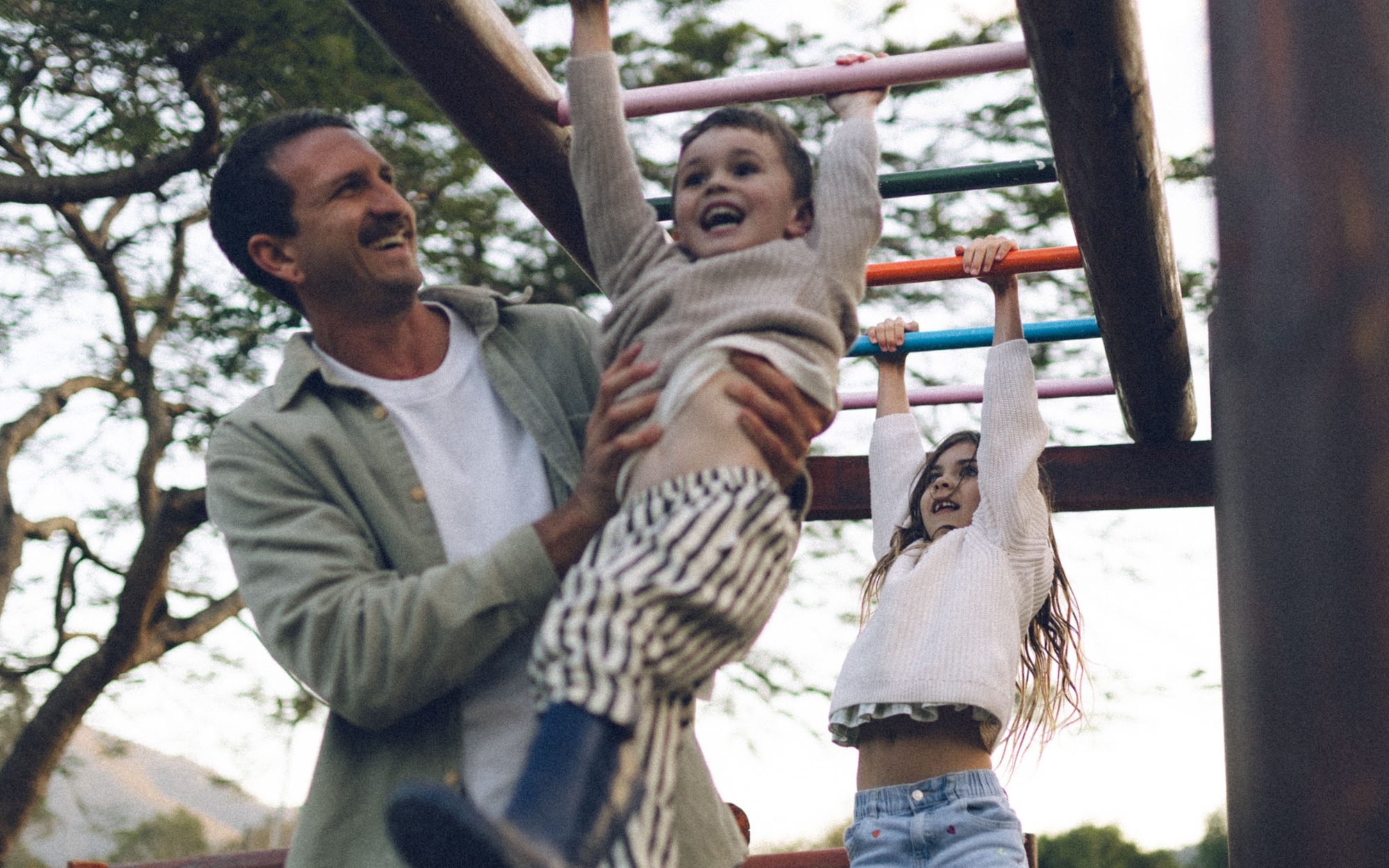 A father laughing as he lifts his son to the monkey bars while his daughter swings along beside them at the Historic Rivermill playground