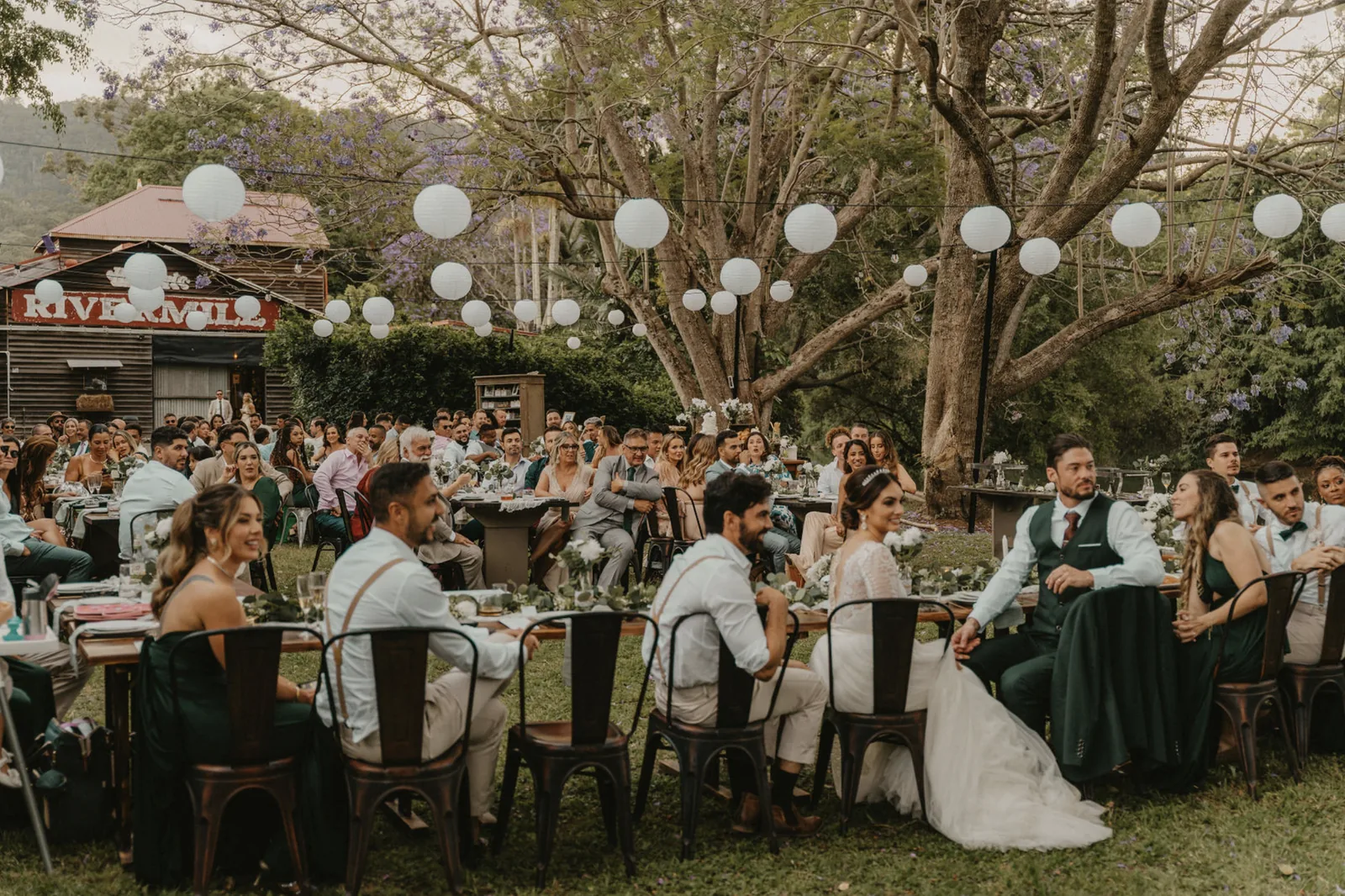 Guests seated at long tables under paper lanterns in the jacaranda gardens at Historic Rivermill