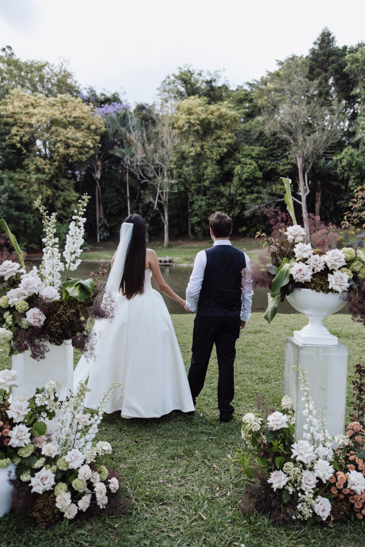 A couple hand in hand between floral arrangements, looking to the Coomera River