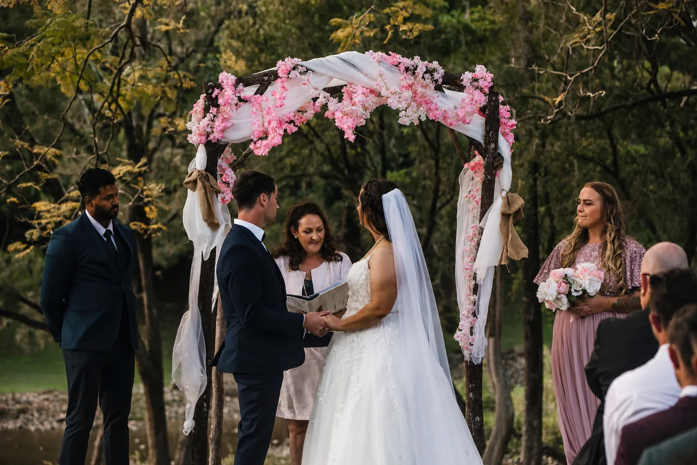 A couple holding hands with their celebrant beneath a blossom-draped timber arbour by the Coomera River