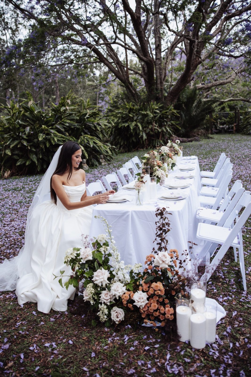 A bride at a long white reception table under a jacaranda in bloom, petals carpeting the lawn